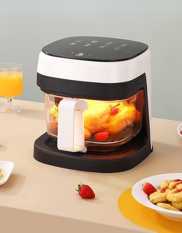 Black and white air fryer with food inside on a table with a glass of orange juice and a bowl of fruit. Transparent 8.5L glass air fryer with smart touch panel cooking a whole chicken.